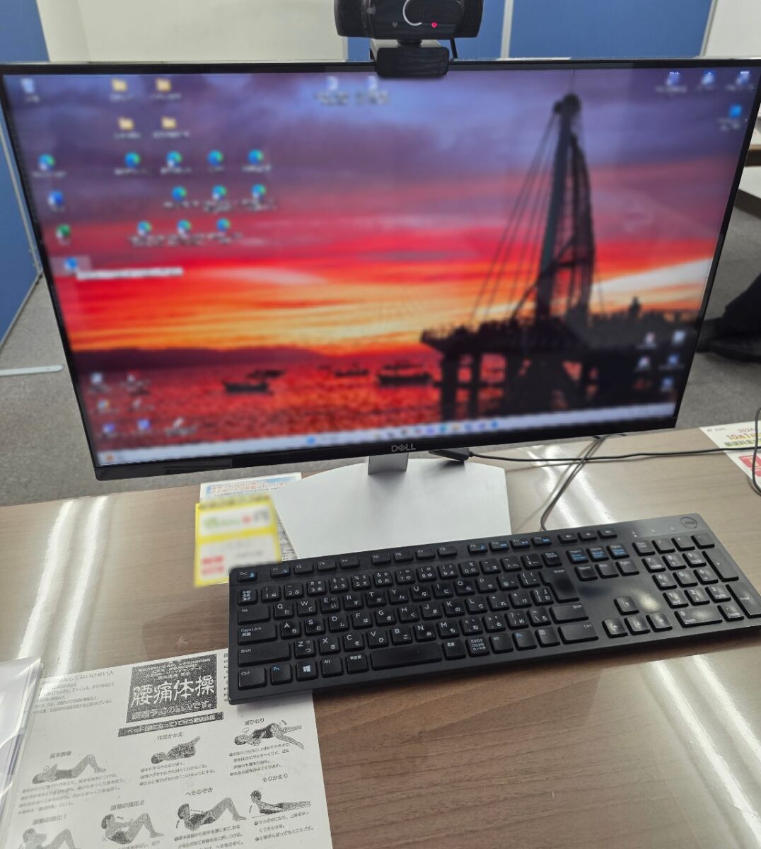Dell製モニターとワイヤレスキーボードが設置されたデスク。モニターには夕焼け空とクレーンの風景の壁紙が表示され、ウェブカメラがモニター上部に置かれている。