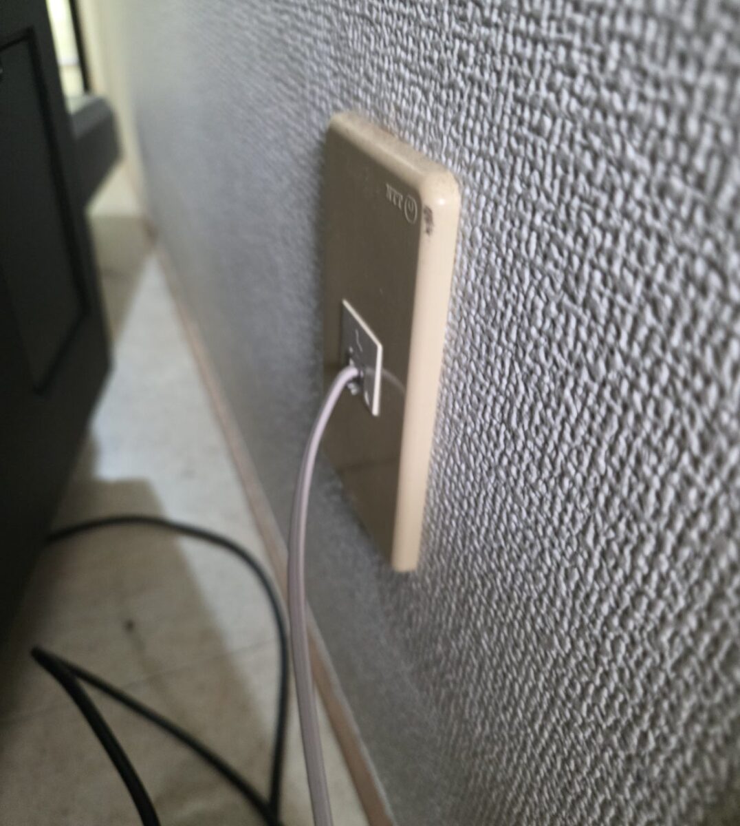 壁に設置されたNTTロゴ入りのモジュラージャックに電話線が接続されている様子。